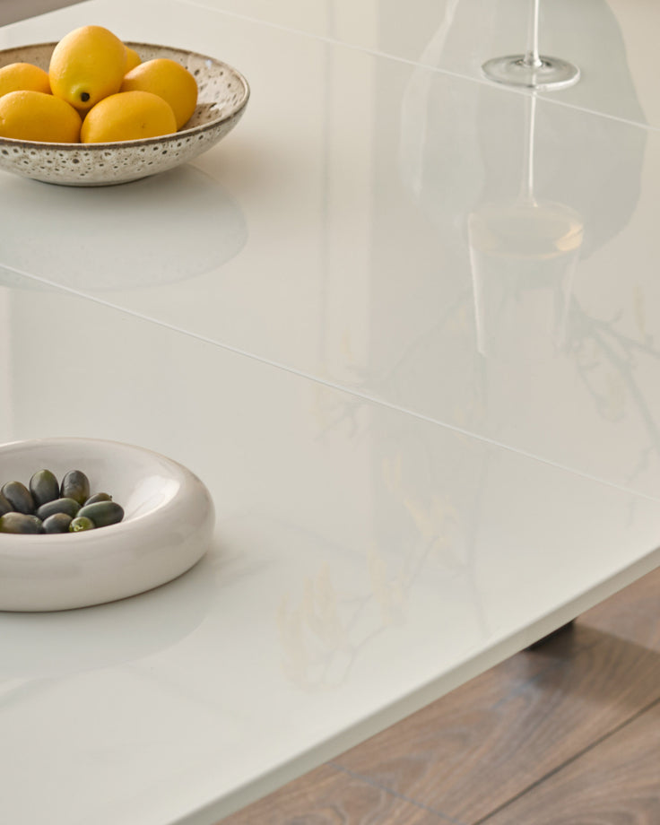 White extending dining table with glossy finish, styled with bowls of lemons and olives, wooden base visible.