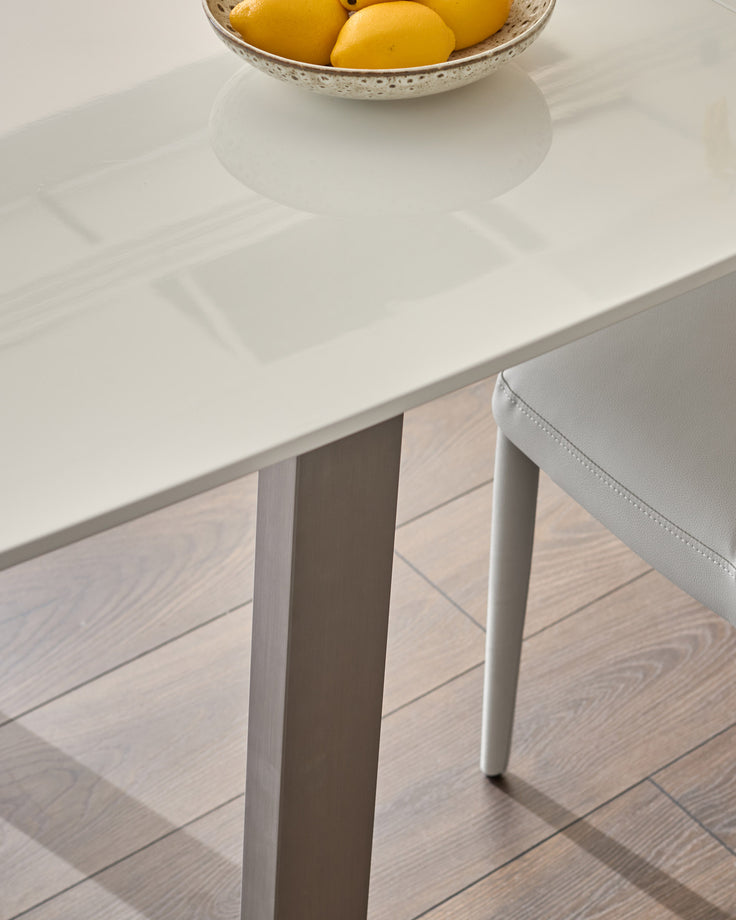 White extending dining table with clean lines, shown at an angle to display its slender legs and minimalist design on light wood flooring.