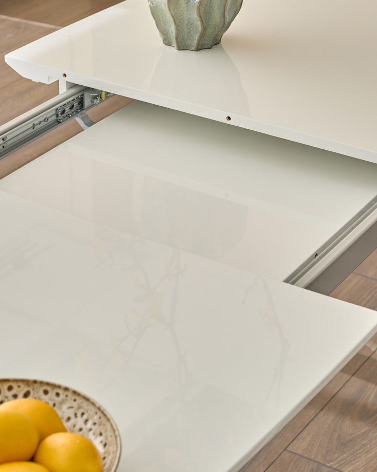 White extending dining table with open drawer, showing metal slide mechanism and glossy finish, styled with cactus and lemons.