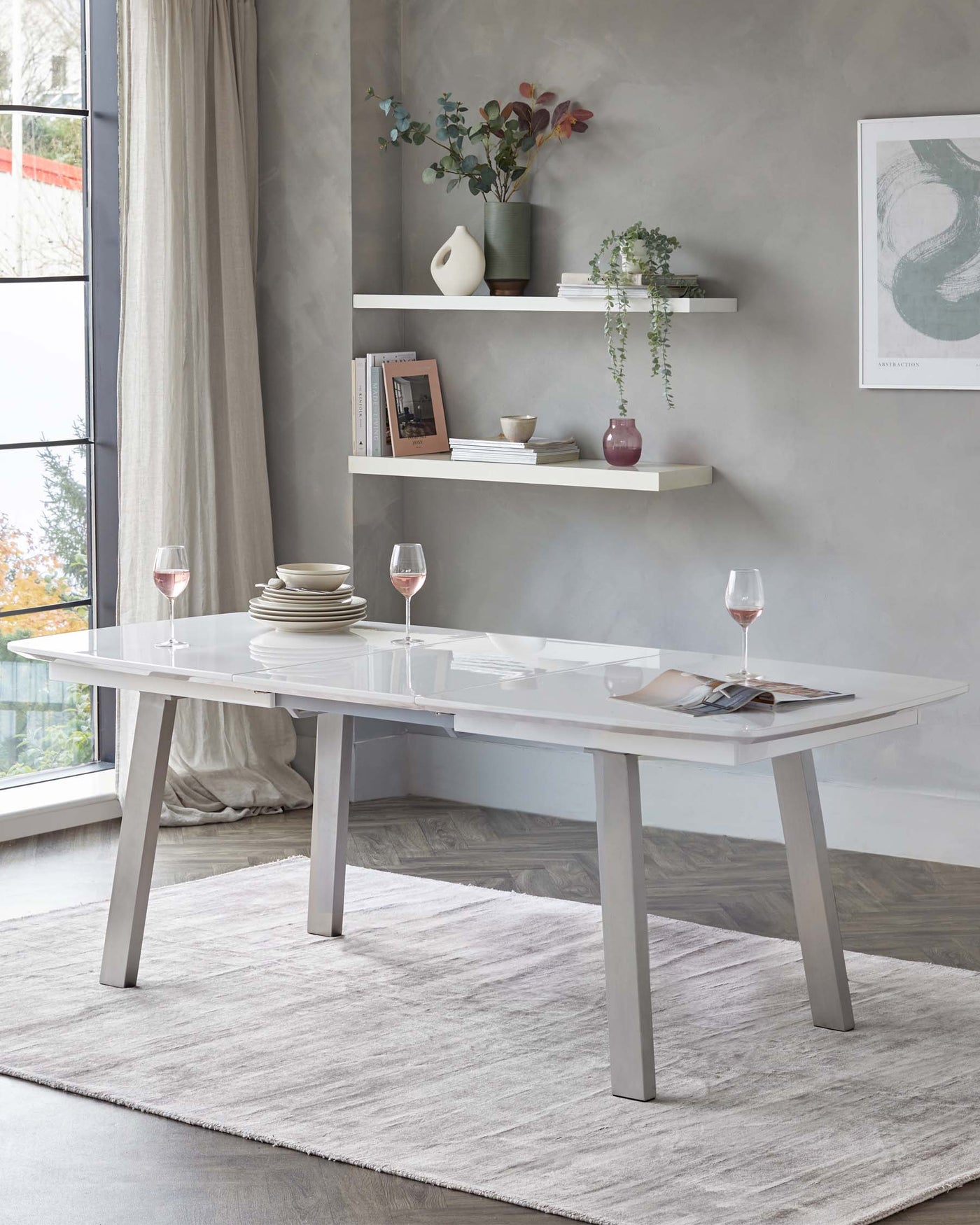 Modern light grey dining table with a reflective surface and angled legs, accompanied by two white floating shelves on a textured wall above, displaying decorative items and books. A soft grey area rug lies beneath the table.