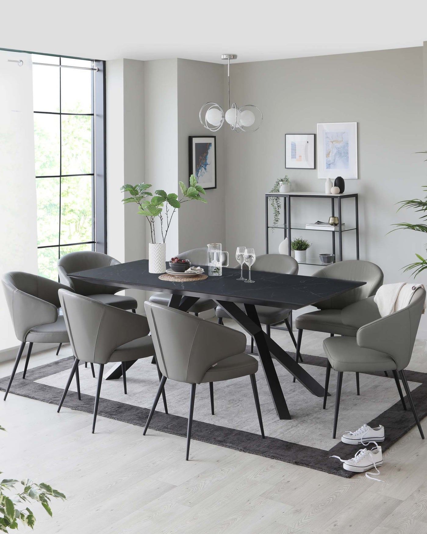 Contemporary dining room featuring an oval-shaped, dark wood table with a matte finish and angular black legs. Six matching upholstered chairs with a sleek design in a muted grey tone, complete with slender black metal legs, are positioned around the table. A minimalist black metal shelving unit displaying decorative objects provides a functional backdrop. The clean lines and neutral colour palette create a modern yet inviting atmosphere.