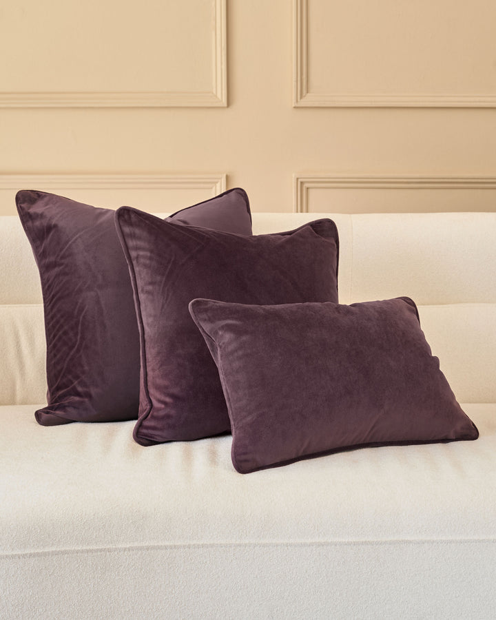 Luca Cushion in mulberry velvet, displayed as a set of three on a cream upholstered bed against a paneled wall.