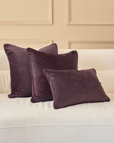 Luca cushion in mulberry velvet, displayed in a set of three on a cream sofa against a paneled cream wall.