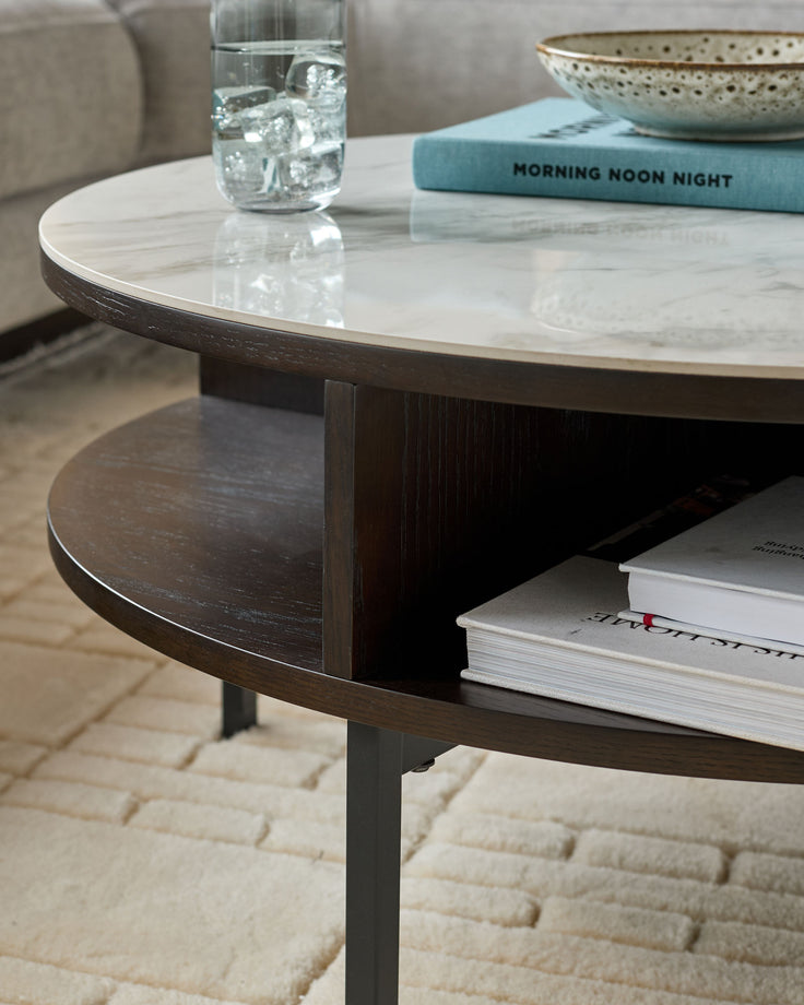 Aurora round coffee table with white marble top, dark oak base, and lower shelf, styled with water glass and books.