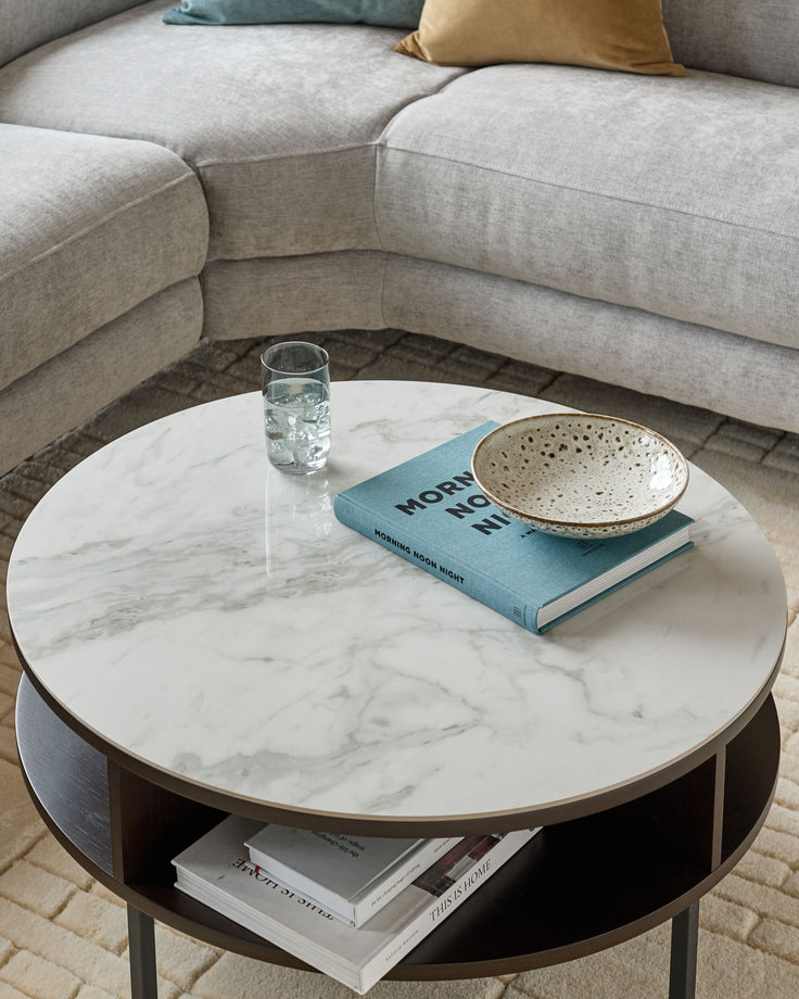 Aurora round marble coffee table with white marble top, oak base, and black open shelf storage, styled in living room setting.