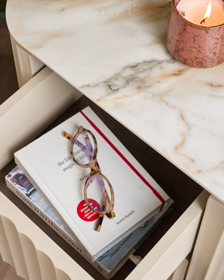 Austin bedside table in natural ceramic finish, styled with books, glasses, and a lit candle on marble top.
