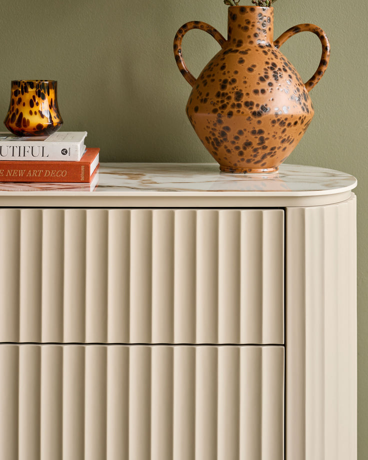 Austin chest of drawers in natural ceramic with vertical fluted detailing and rounded marble top, styled with decorative objects.