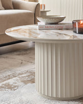 A round coffee table with marble top and fluted base sits on a cream and beige patterned rug. A bowl, book and candle sit on top.