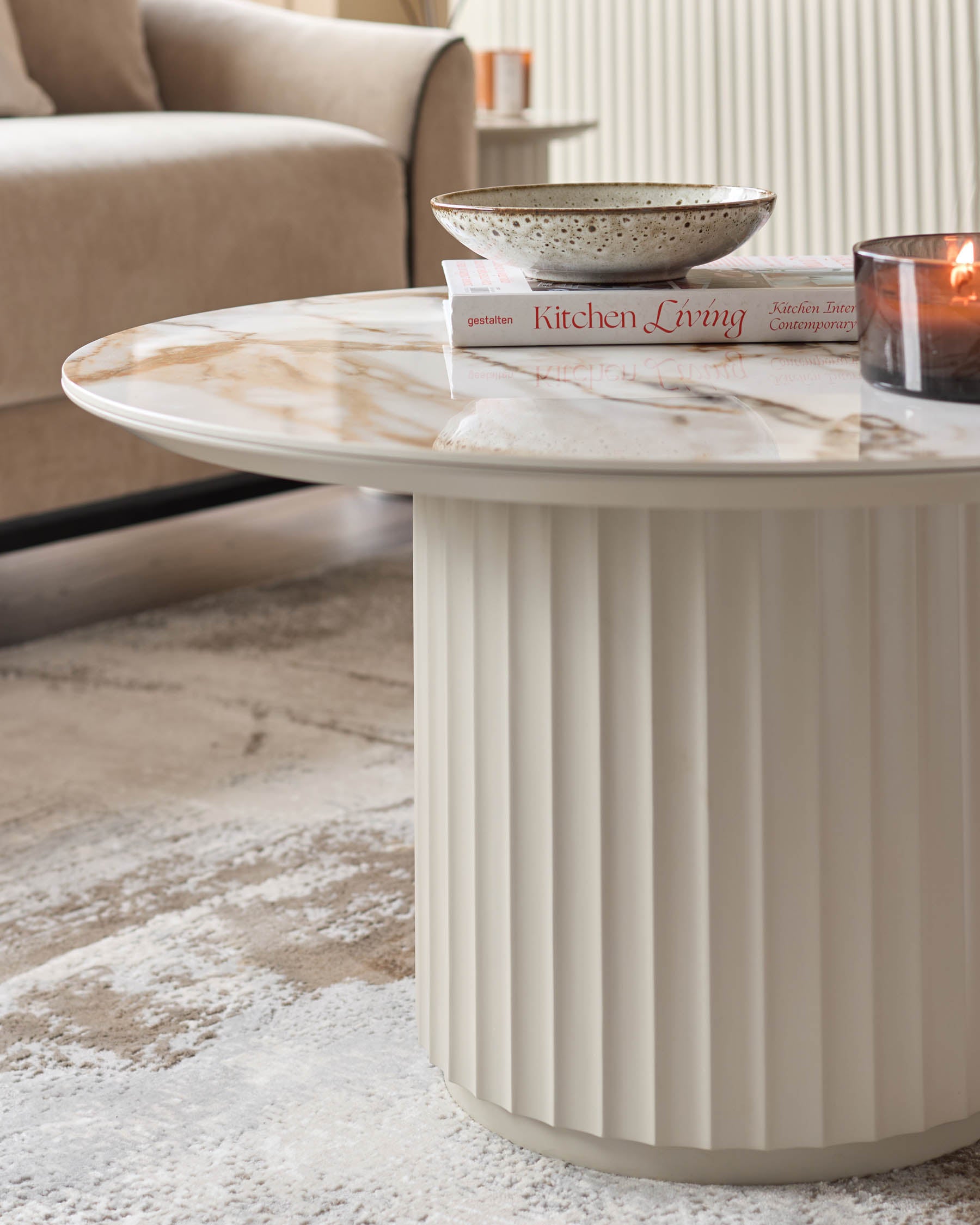 A round coffee table with marble top and fluted base sits on a cream and beige patterned rug. A bowl, book and candle sit on top.