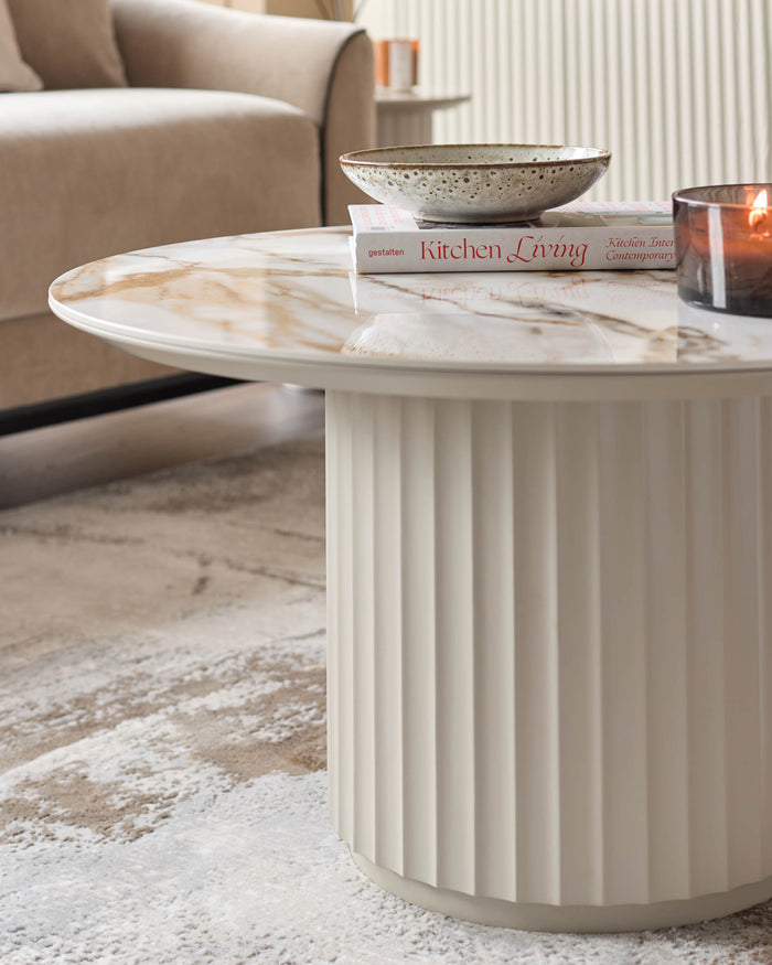 A round coffee table with marble top and fluted base sits on a cream and beige patterned rug. A bowl, book and candle sit on top.