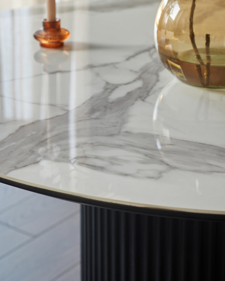 A round marble table with a ribbed black base, featuring a candle holder and a vase with branches on top.