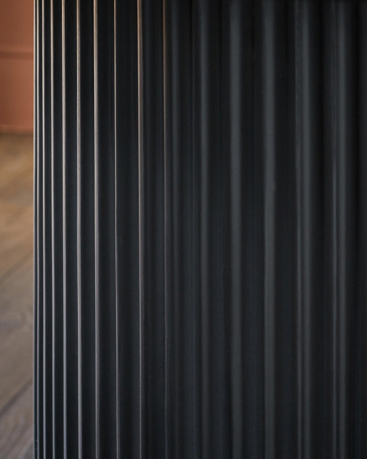 Close-up of a fluted, dark grey, metal table leg. A glimpse of the wooden floor and a pink wall can be seen in the background.