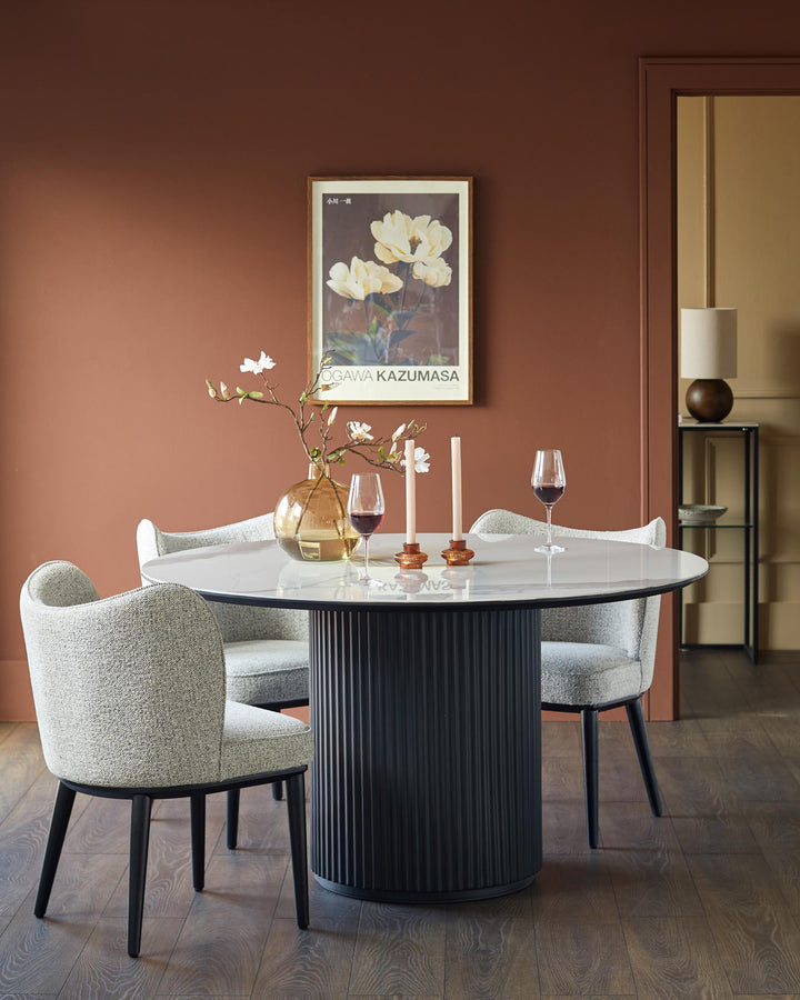 A round dining table with a grey marble top and a dark ribbed base is surrounded by four upholstered chairs with black legs.