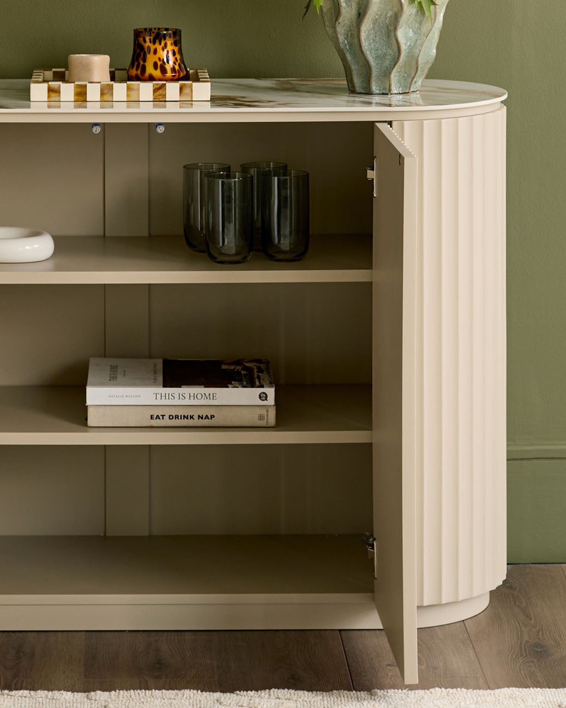 Austin compact sideboard in natural ceramic finish, curved design with open shelving displaying glassware and books.