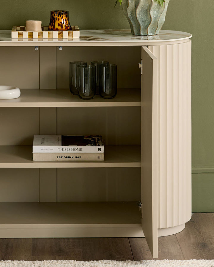 Austin compact sideboard in natural ceramic finish, curved design with open shelving displaying glassware and books.