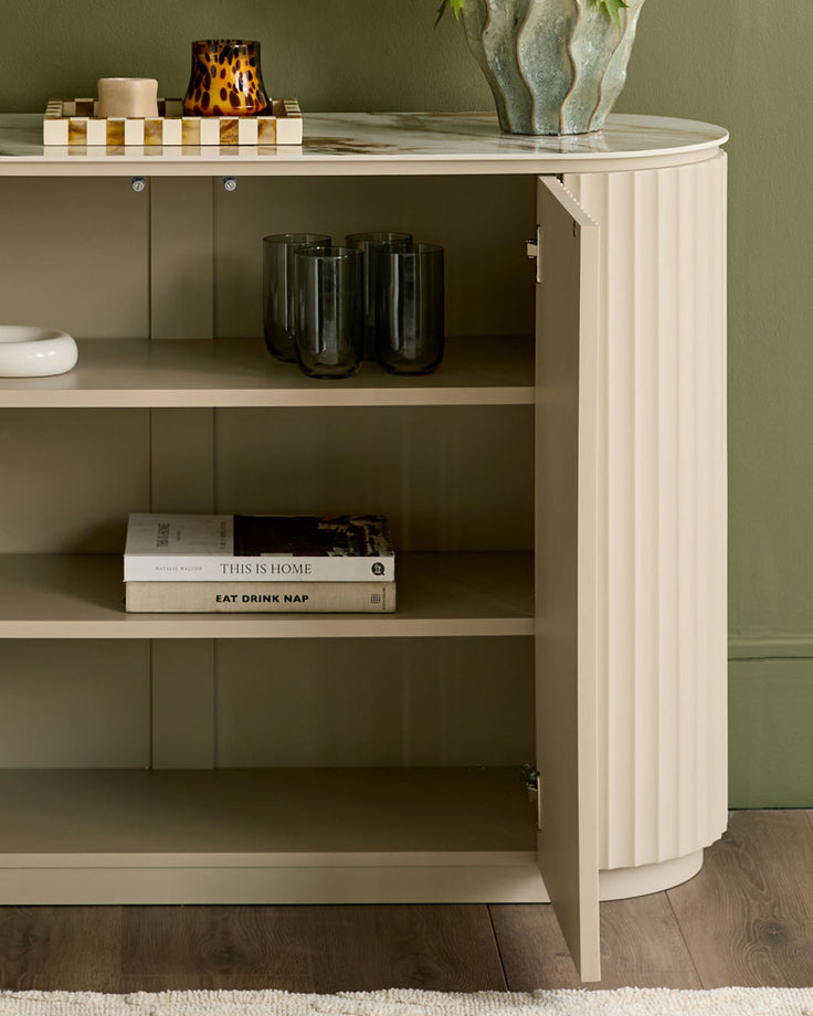 Austin compact sideboard in natural ceramic finish, curved design with open shelving displaying glassware and books.