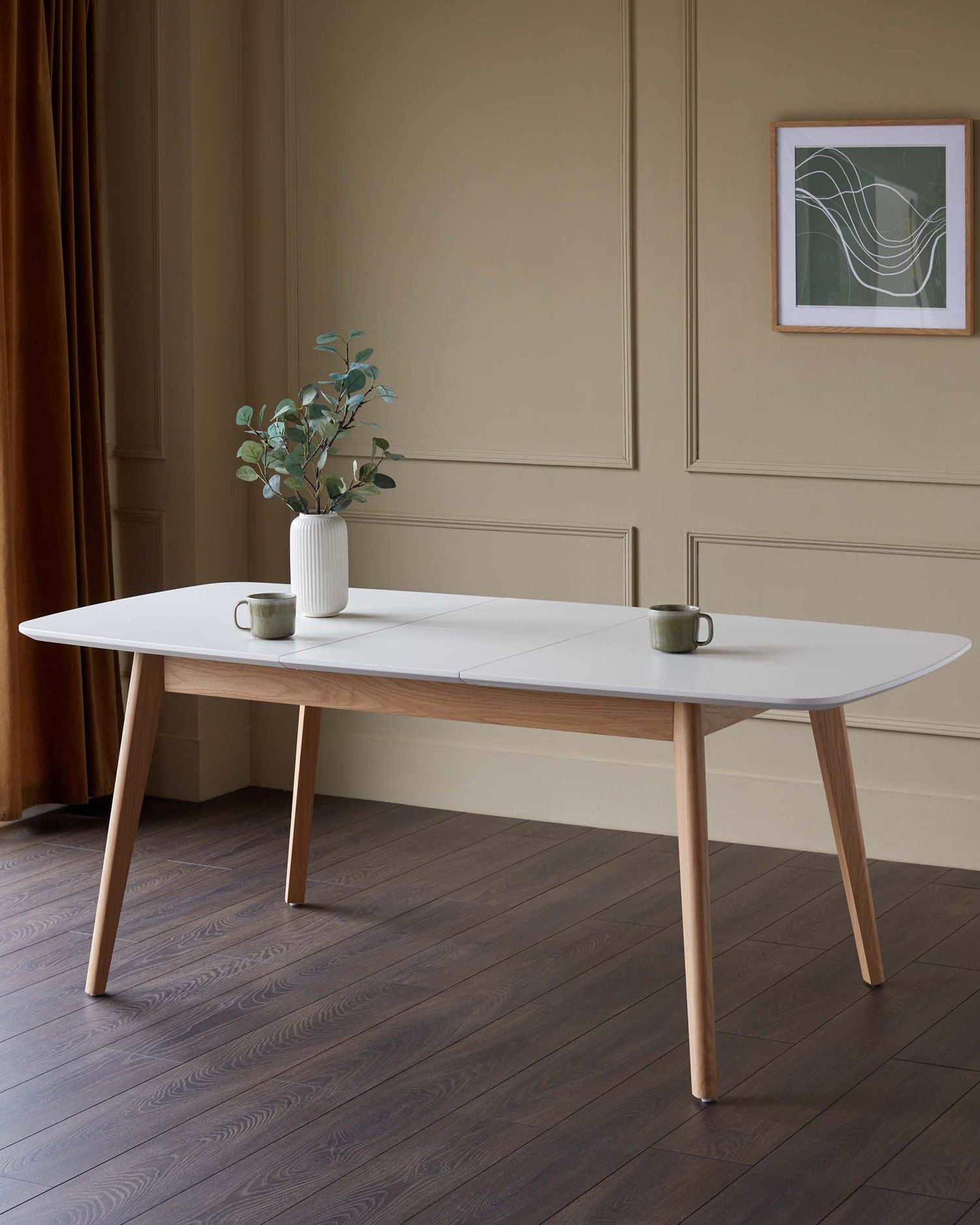 A modern dining table with a white top and wooden legs, styled with cups and a vase of foliage.