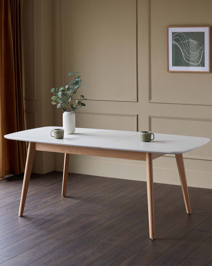A modern dining table with a white top and wooden legs, styled with cups and a vase of foliage.