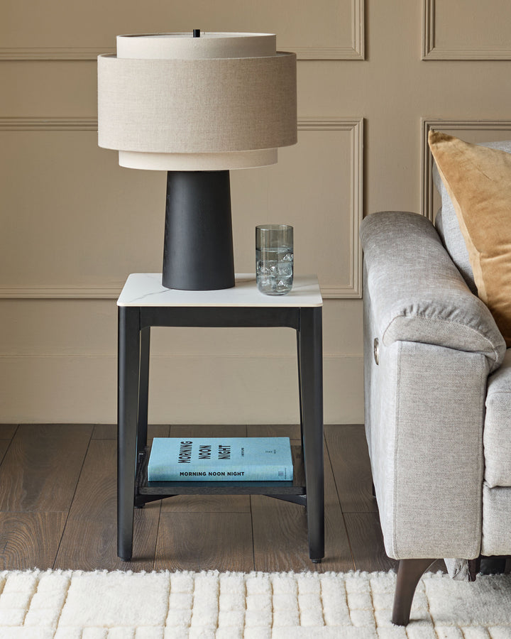Leonie table lamp with natural linen shade and mocha base, displayed on black side table next to Grey sofa.
