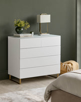White chest of drawers with four drawers and gold legs, styled with a lamp, vase and books.