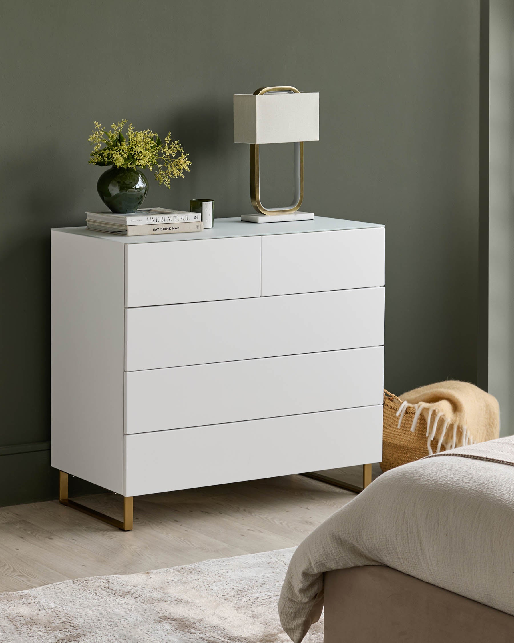 White chest of drawers with four drawers and gold legs, styled with a lamp, vase and books.