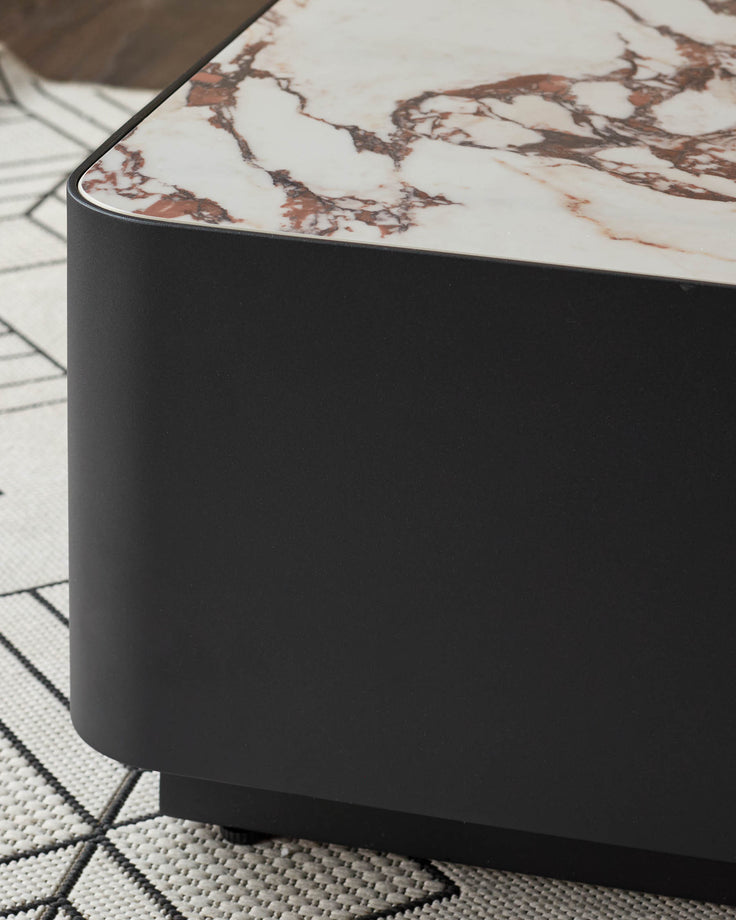 Black cylindrical coffee table with white Calacatta marble top featuring brown veining, shown from a side angle on a geometric patterned rug.