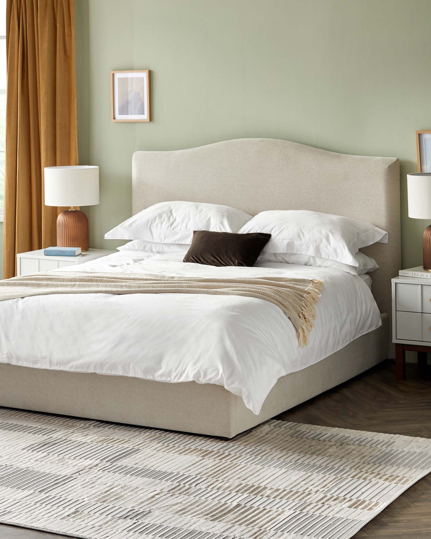 A contemporary bedroom scene featuring a large upholstered bed with a curved headboard in a neutral colour, dressed with crisp white bedding accented by a dark throw pillow. Flanking the bed are two matching white nightstands with a modern design, each adorned with a table lamp featuring a cylindrical shade and a warm-toned base. The room's decor is completed with a textured striped area rug on a wood floor, adding depth and interest to the space.