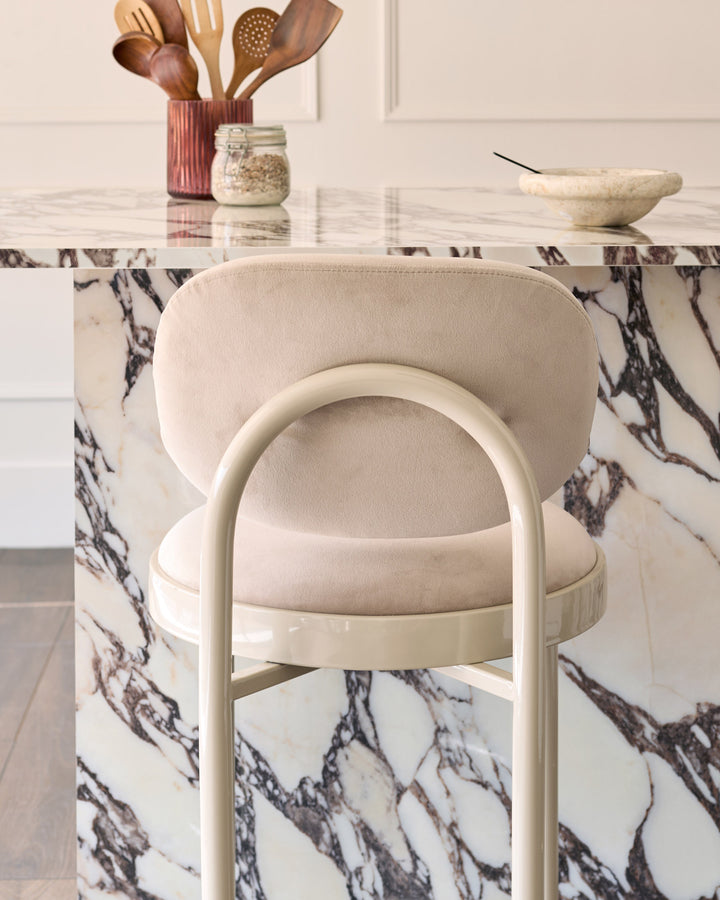 Blake bar stool in champagne velvet with curved backrest and cream metal frame, shown at marble table.