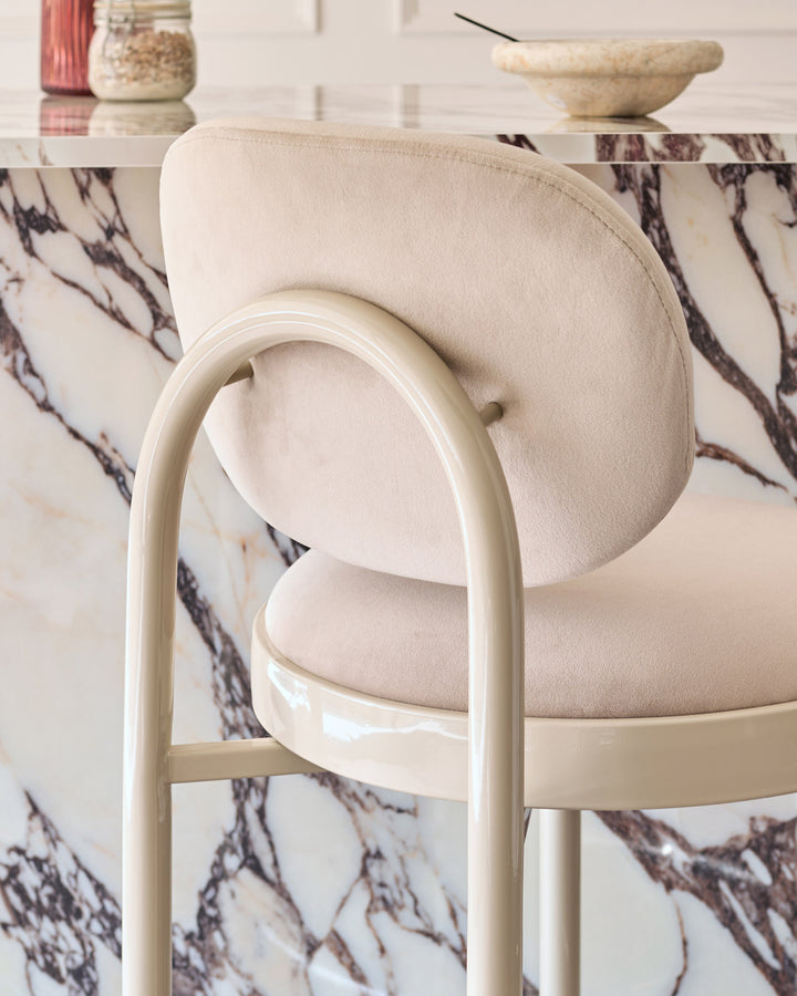Blake bar stool in champagne velvet with cream curved frame, shown at close angle against marble backdrop.