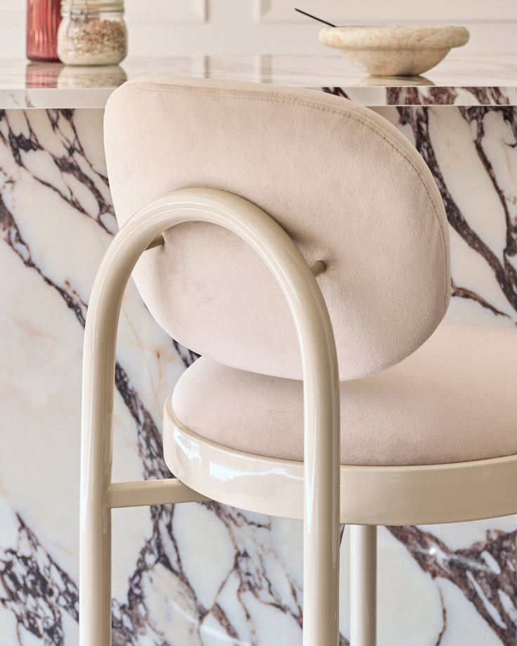Blake bar stool in champagne velvet with cream curved frame, shown at close angle against marble backdrop.