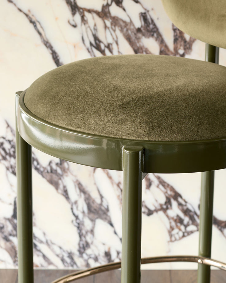 Blake bar stool in olive green velvet with polished metal frame, shown at an angle against marble wallpaper backdrop.