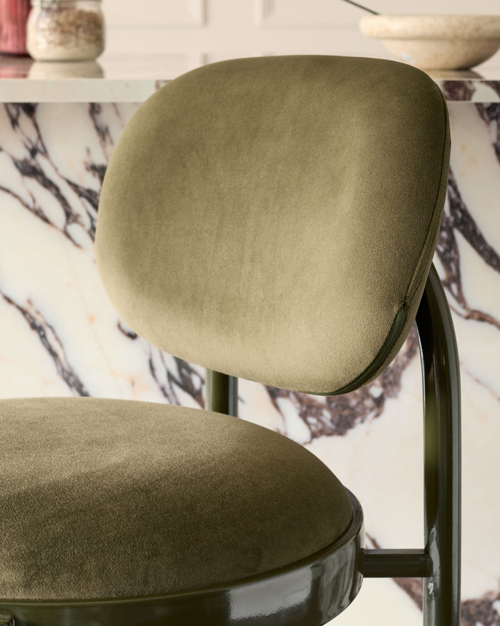 Blake bar stool in olive green velvet with curved backrest and metal frame, shown from side angle.