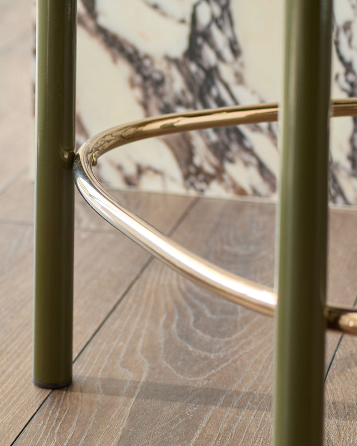 Blake bar stool in olive green velvet with brass footrest detail, close-up side view on wood flooring.