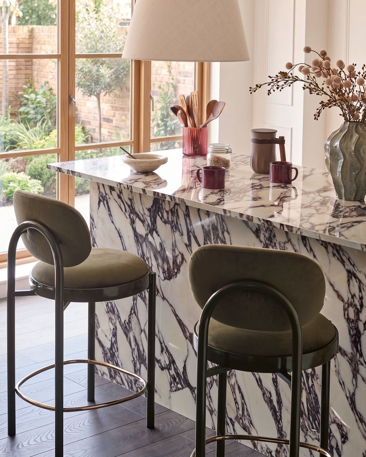 Blake bar stool in olive green velvet with curved backrest and metal frame, shown at marble counter.