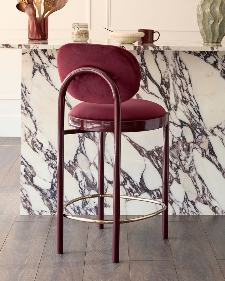 Blake bar stool in plum velvet with burgundy metal frame and brass footrest, shown at marble-topped counter.