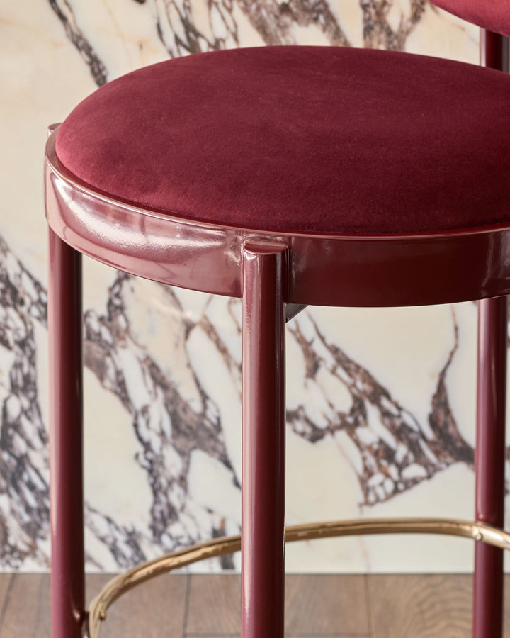Blake bar stool in plum velvet with polished brass footrest, shown from side angle against marble wallpaper backdrop.