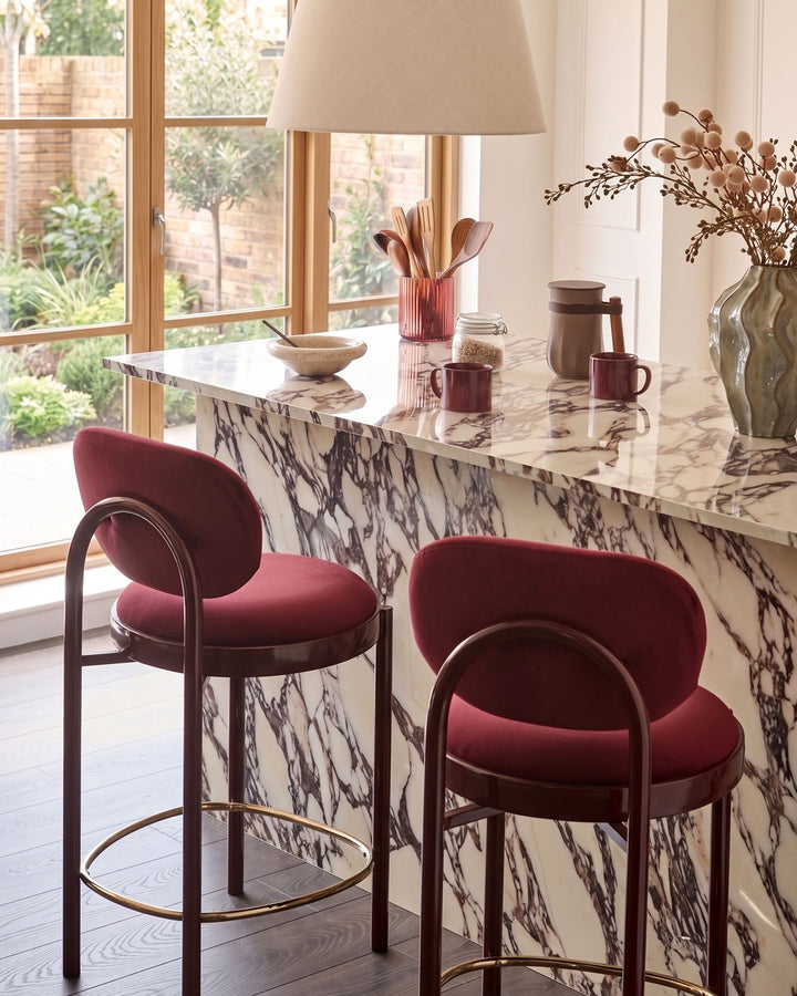 Blake bar stool in plum velvet with curved backrest and bronze metal frame, shown at marble countertop.