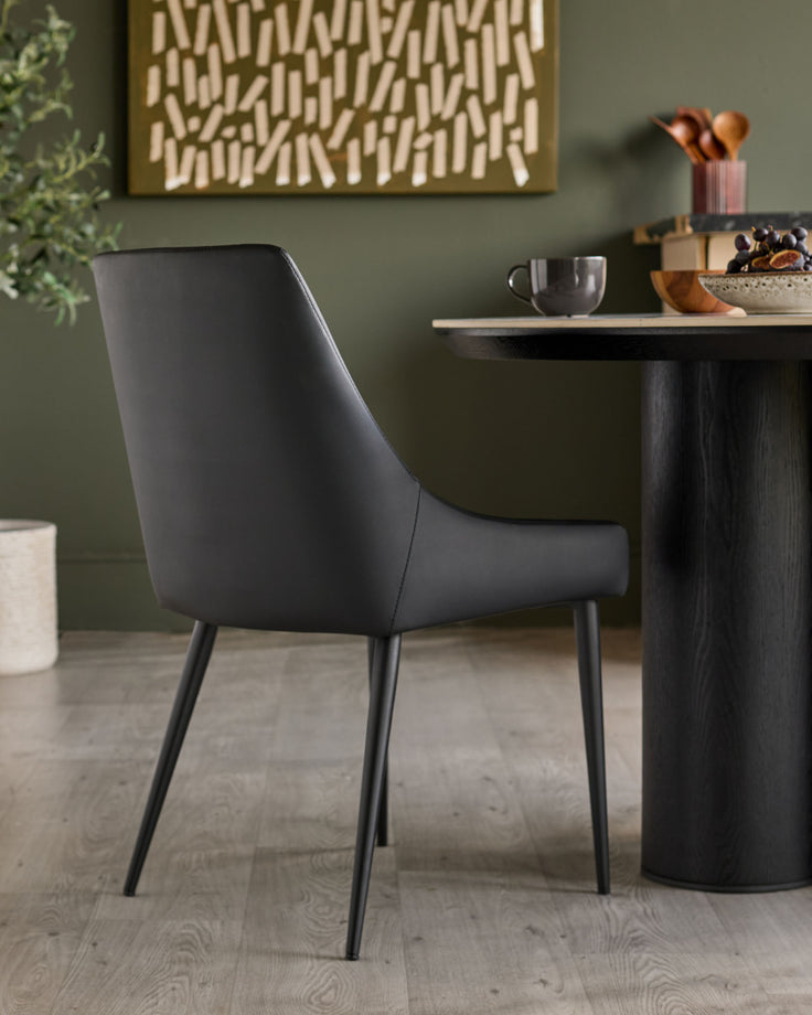 Blakely black faux-leather dining chair with tapered legs, shown in a styled room setting beside a dark wooden table.