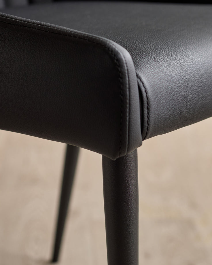 Close-up of black faux leather dining chair showing textured seat padding and dark metal leg detail.