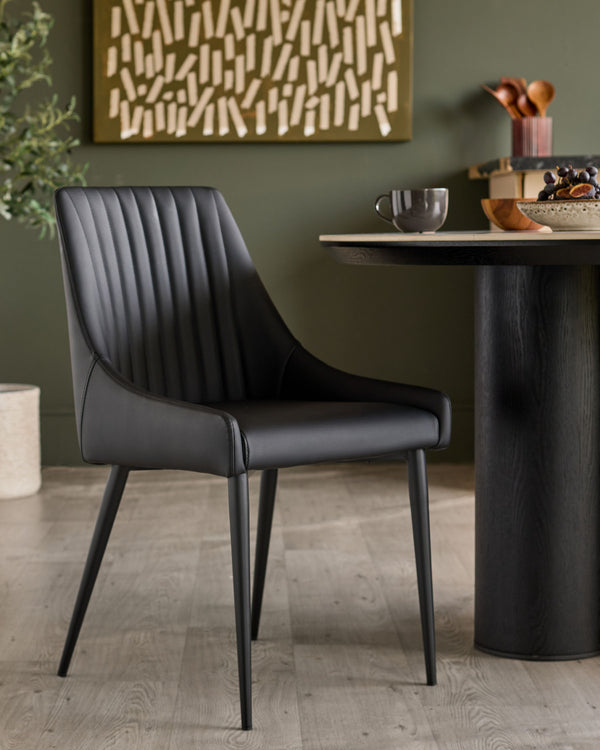 Black faux leather dining chair with vertical stitched back, curved seat, and tapered metal legs, shown in room setting.