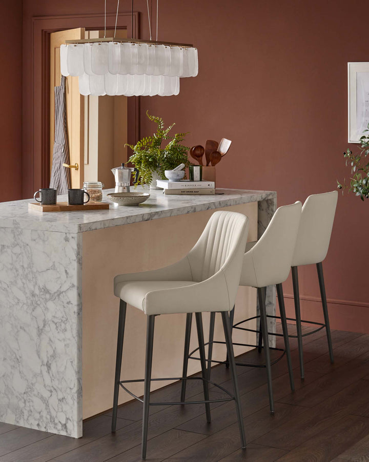 A kitchen island with marble countertop and two cream bar stools with black legs, under a layered glass chandelier.