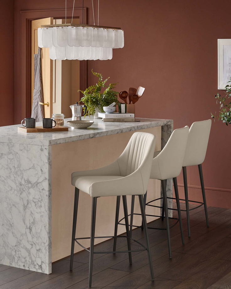 A kitchen island with marble countertop and two cream bar stools with black legs, under a layered glass chandelier.