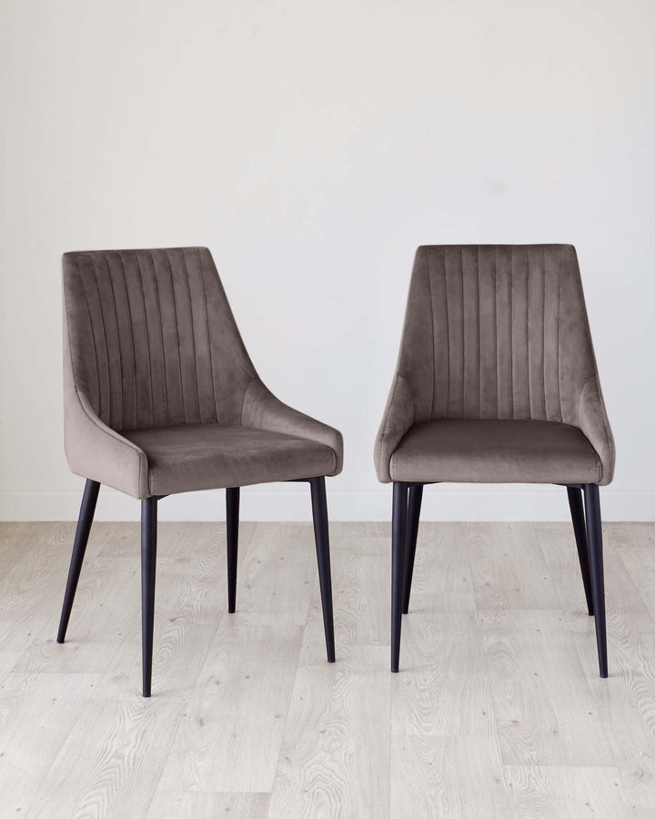 Two modern dining chairs upholstered in taupe velvet with black legs, standing on a light wooden floor against a white wall.