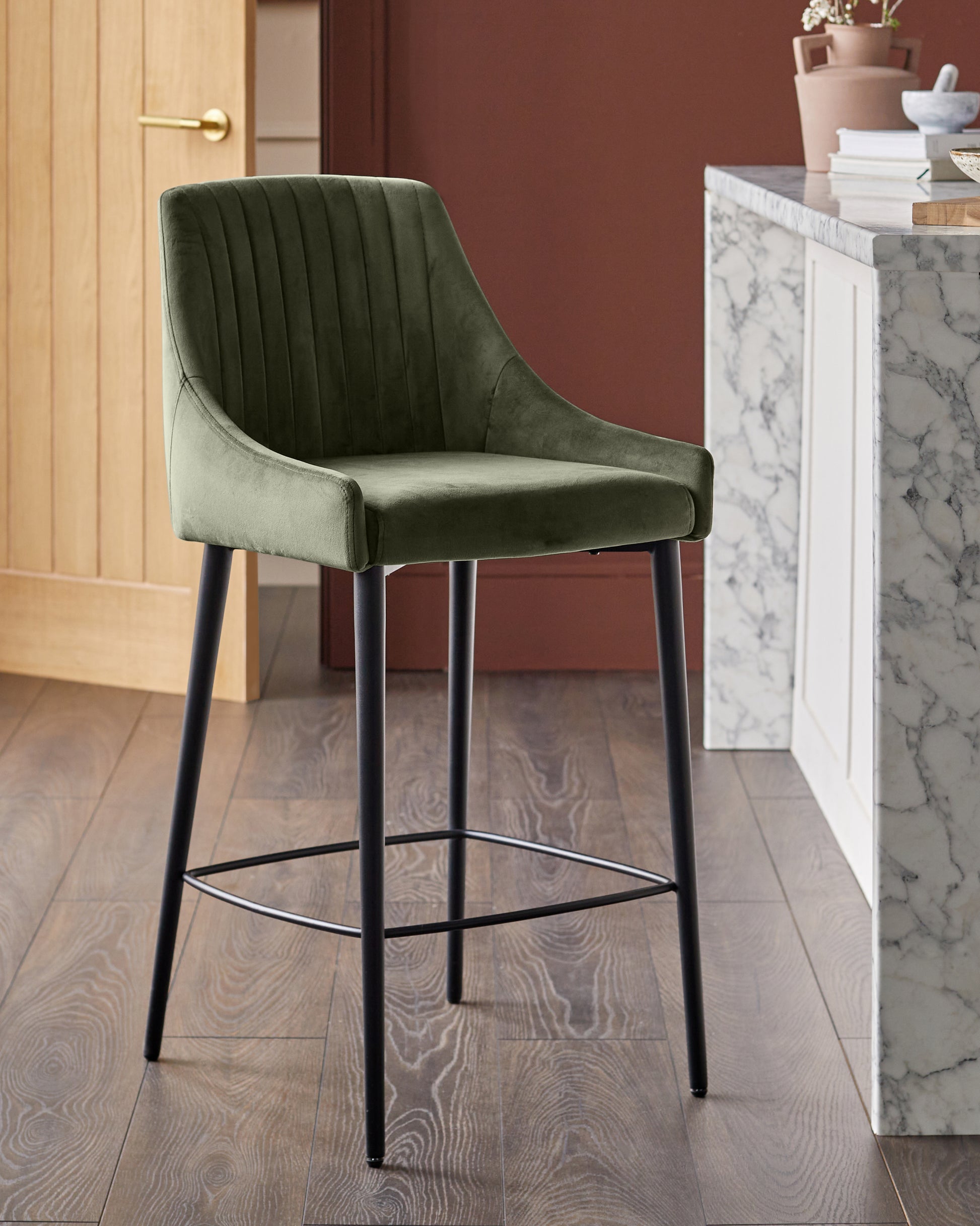 Blakely bar stool in olive green velvet with vertical stitching, black metal legs and footrest.