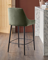 Blakely bar stool in olive green velvet with black metal legs and footrest, shown in modern kitchen setting.