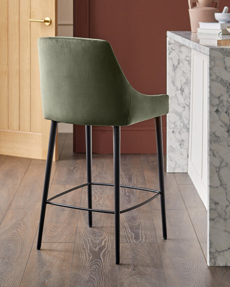 Blakely bar stool in olive green velvet with black metal legs and footrest, shown in modern kitchen setting.