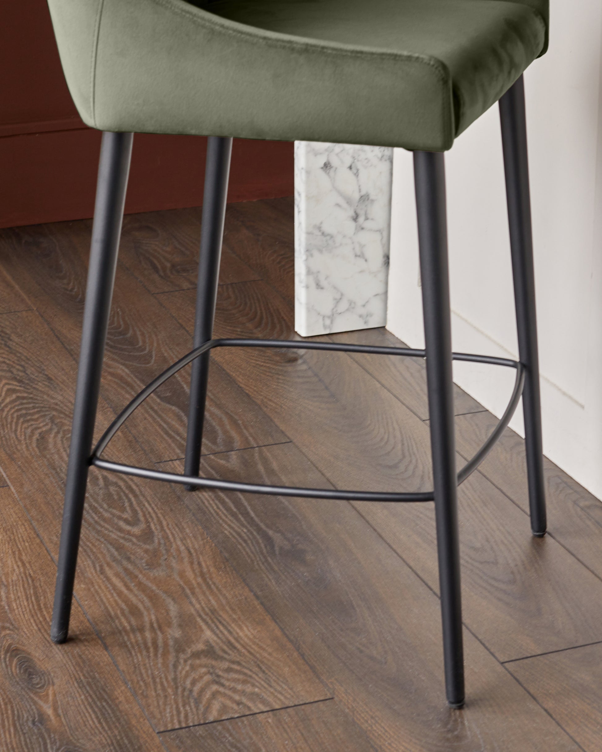 Blakely bar stool in olive green velvet with black metal legs and footrest, shown from side angle on wood flooring.