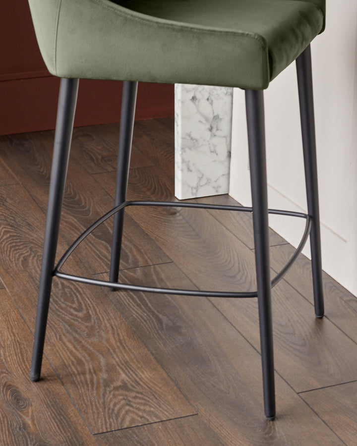 Blakely bar stool in olive green velvet with black metal legs and footrest, shown from side angle on wood flooring.