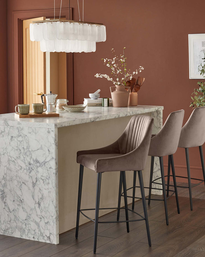 Three modern velvet bar stools sit at a marble-topped kitchen island under a stylish layered pendant light.