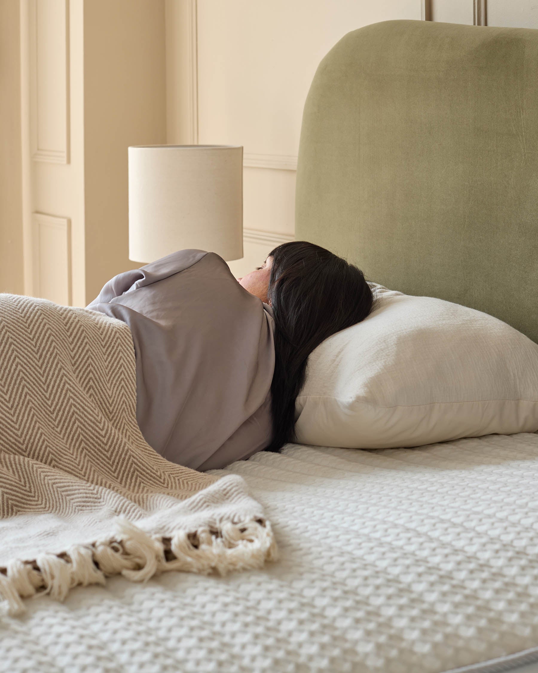 A bedroom setting with a bed, lamp and headboard. The bed is covered with a textured throw and a woman is sleeping on it.